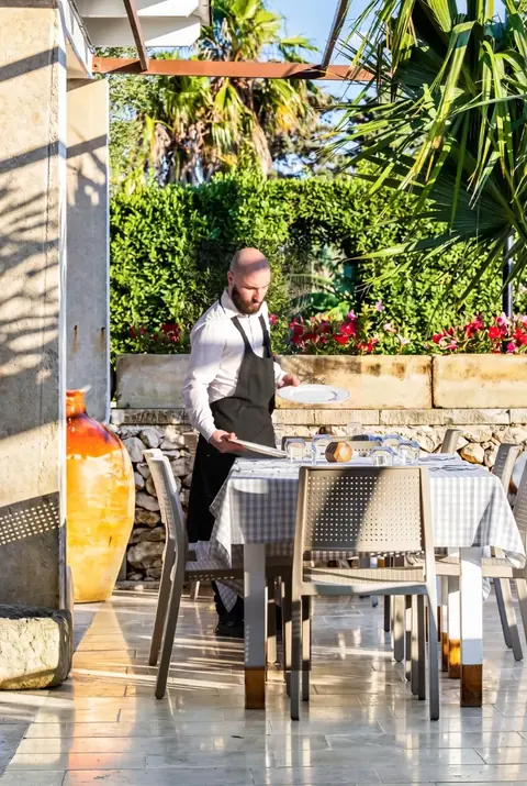 Un cameriere apparecchia i tavoli sotto il portico, con il giardino alle spalle